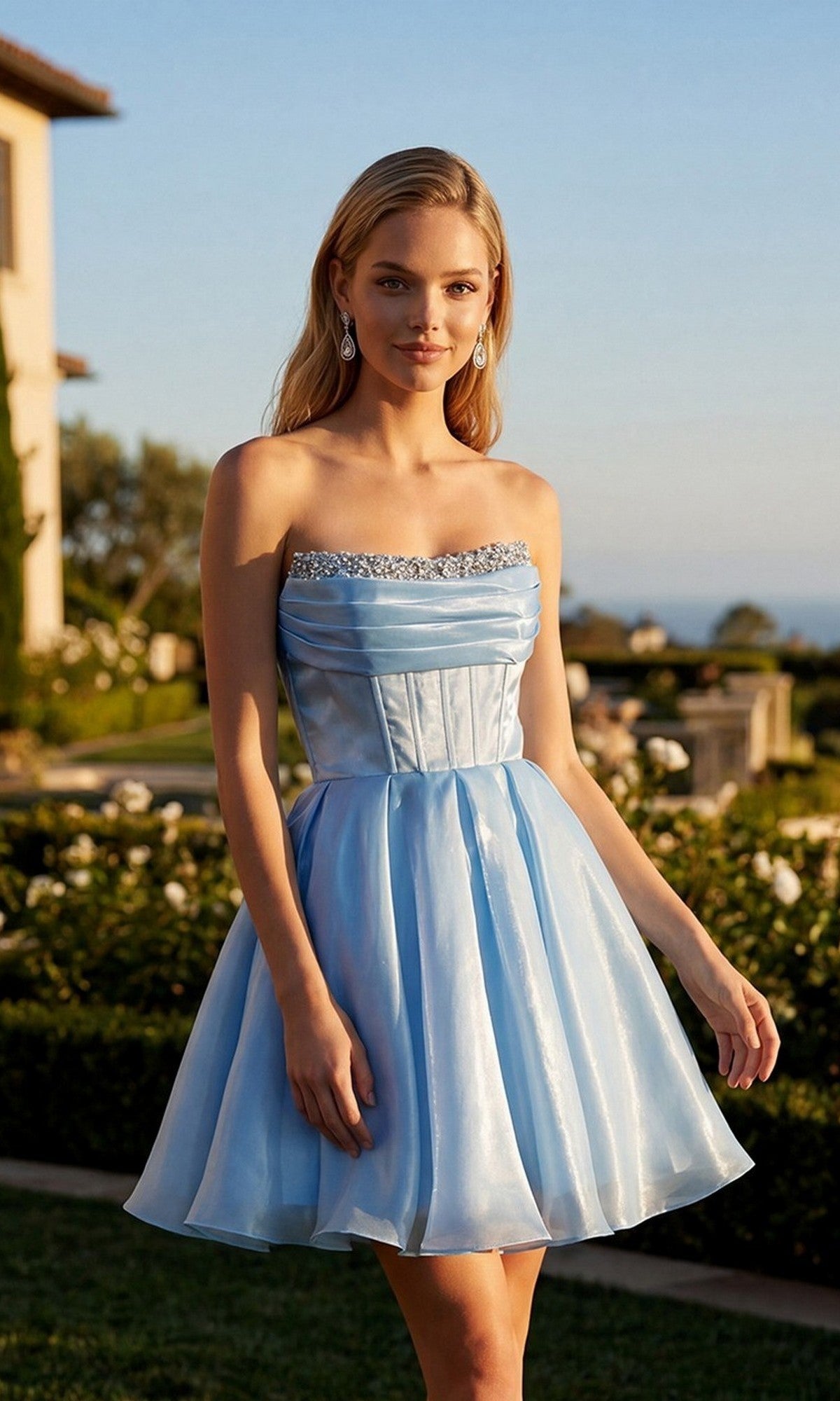 A woman stands outdoors wearing the Short Homecoming Dress: Juliet JT7187S in Sky Blue, which features a strapless design with a fitted corset bodice and beaded neckline. The knee-length shimmer organza skirt catches the light beautifully, set against a backdrop of lush greenery and a charming building.